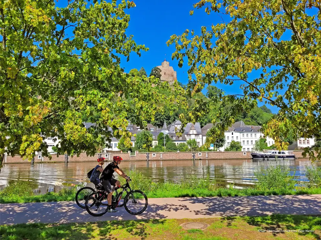 Zwei Menschen fahren mit dem Fahrrad auf einem von grünen Bäumen gesäumten Uferweg entlang und genießen die schöne Umgebung. Unter einem strahlend blauen Himmel sind auf der anderen Seite der Saar weiße Gebäude und eine Burg aus Stein auf einem Hügel zu sehen.