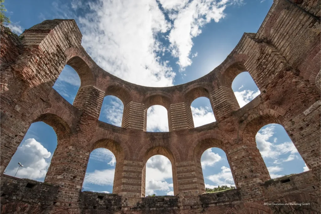 Runde Ziegelruinen mit zahlreichen Bogenfenstern, von unten betrachtet vor einem teilweise bewölkten Himmel an der Saar; Sonnenlicht scheint durch die Öffnungen und beleuchtet die historische Umgebung.